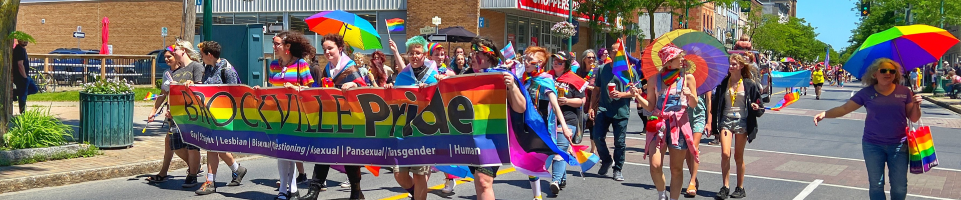 Community supporters march in the annual Pride Parade down king street wearing rainbow items, holding rainbow umbrellas, an carrying a Brockville Pride banner
