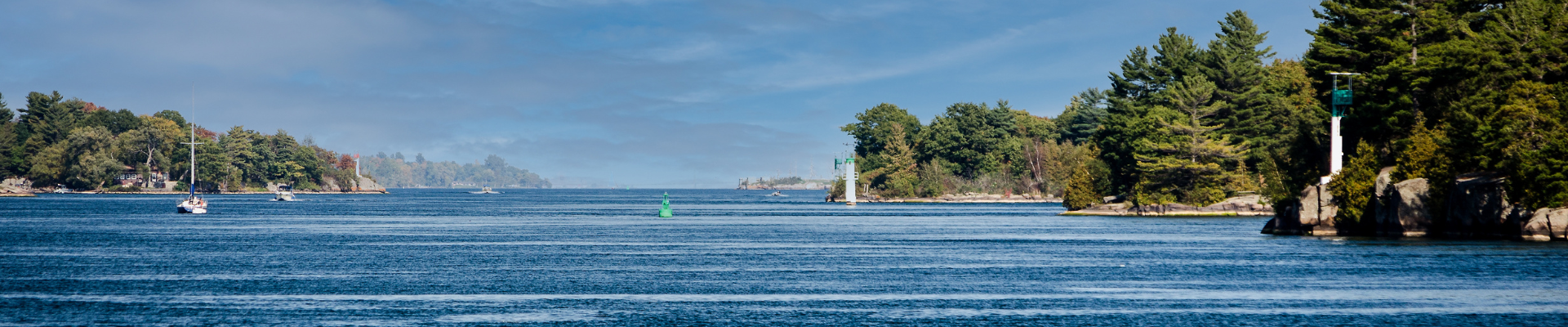 Photo taken looking downriver towards Brockville, between islands in the Saint Lawrence River. Rocky outcroppings jut into the water and host small lighthouses. A green channel marker separates the channel